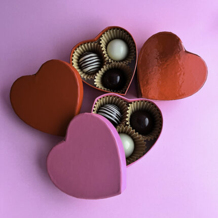 Heart-shaped boxes of artisanal psychedelic bon bons made with Callebaut chocolate, featuring dark, milk, and white chocolate truffles, elegantly arranged on a pink background. A luxurious Valentine's Day gift infused with psilocybin for a unique sensory experience.